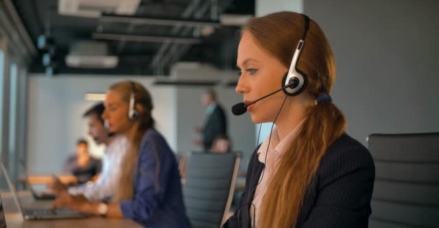 Call-center employees work in deal office. Young handsome female in headset on first plan talking with customer. Modern business room with beautiful view. 4k UltraHD.